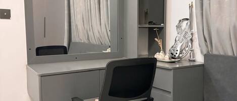 A modern, minimalist bedroom with a grey sofa, a black office chair, a desk with a computer, and a large mirror.