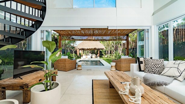 Un salon moderne avec un téléviseur, des meubles en bois, une piscine et une structure dotée d’un toit de chaume.