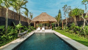 Un espace piscine agrémenté d’un gazebo à toit de chaume, de chaises longues et de plantes tropicales.