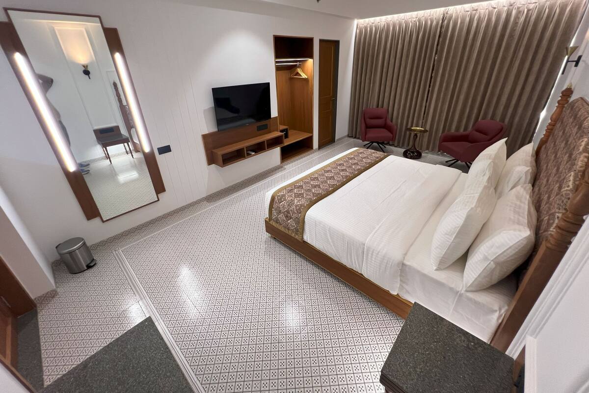 Quarto de hotel com cama grande, televisão, guarda-roupa, área de estar com cadeiras e espelho.