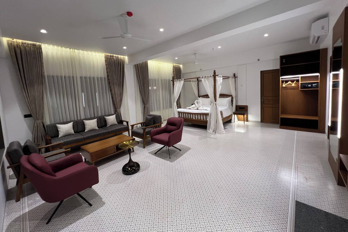 Quarto de hotel moderno com uma cama grande, área de estar com sofá e poltronas, uma mesinha e um guarda-roupa.