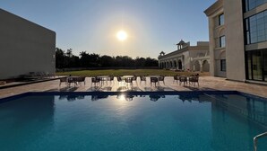 A swimming pool with lounge chairs and tables, surrounded by a building with arched windows and a flat roof.
