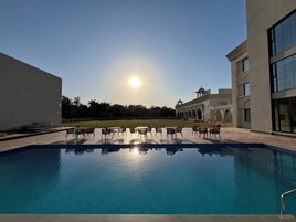 A swimming pool with lounge chairs and tables, surrounded by a building with arched windows and a flat roof.