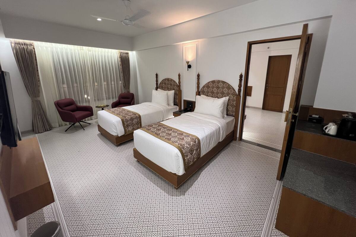 Quarto de hotel com duas camas, uma área de estar com uma poltrona, televisão e um espelho grande.