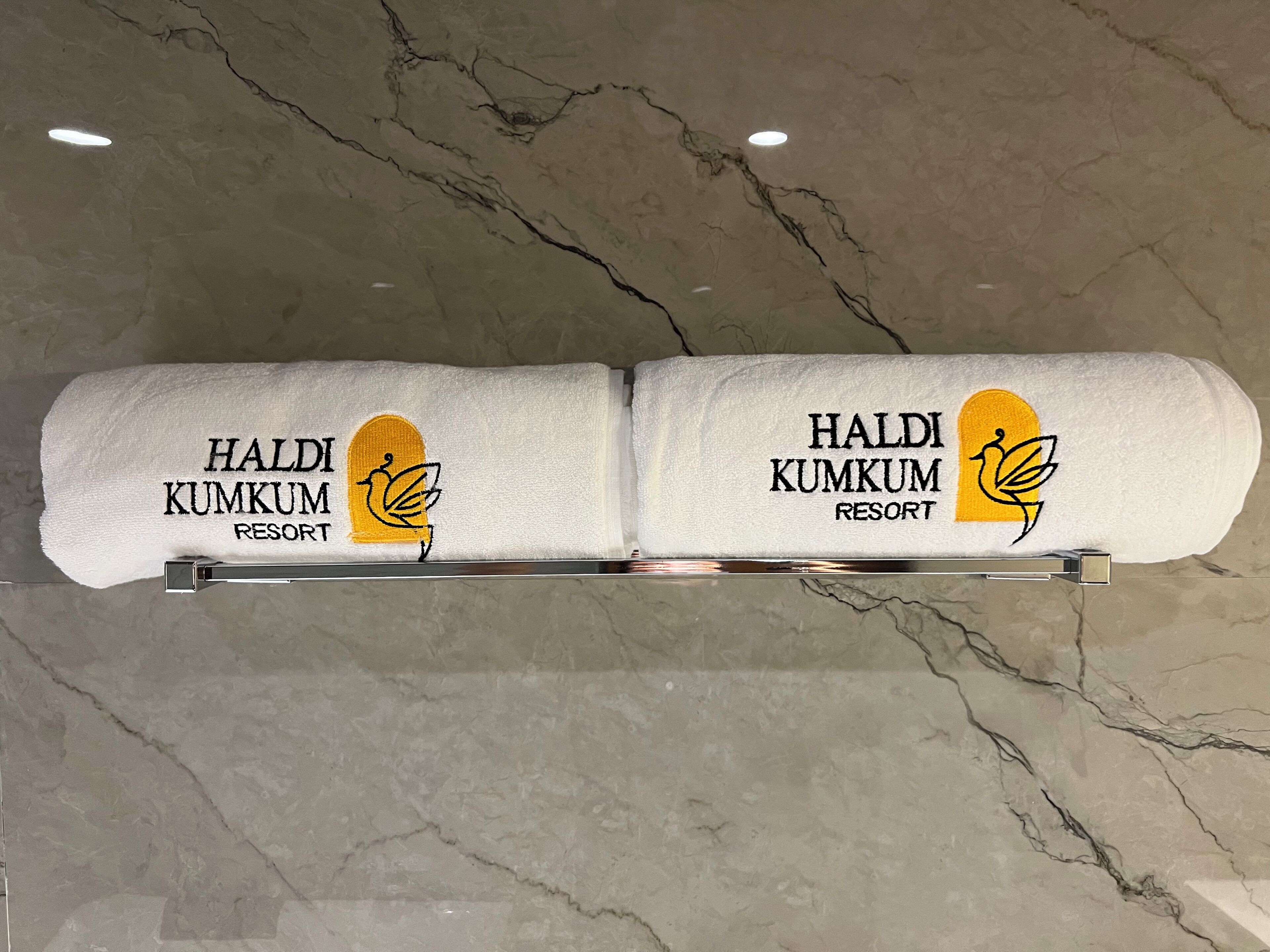 Deux serviettes ornées du texte « HALDI KUMKUM RESORT » et d’un logo sont suspendues à un étendoir en métal, le tout adossé à un mur de marbre.