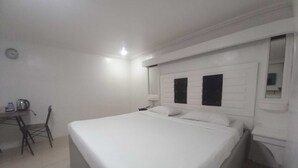 Una habitación sencilla y limpia con una cama, una silla, una mesita con un hervidor y un espejo en la pared.