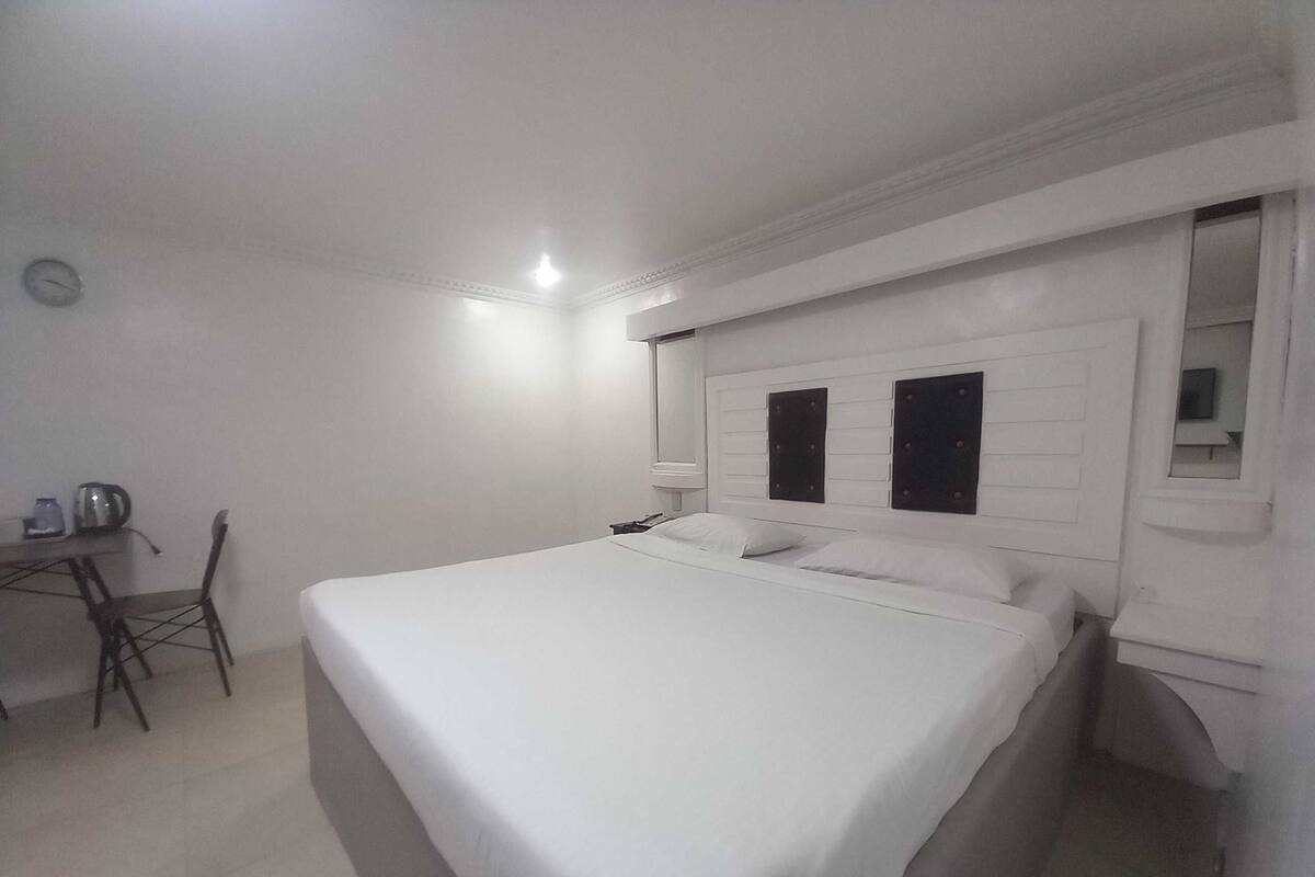 A simple, clean room with a bed, a chair, a small table with a kettle, and a mirror on the wall.