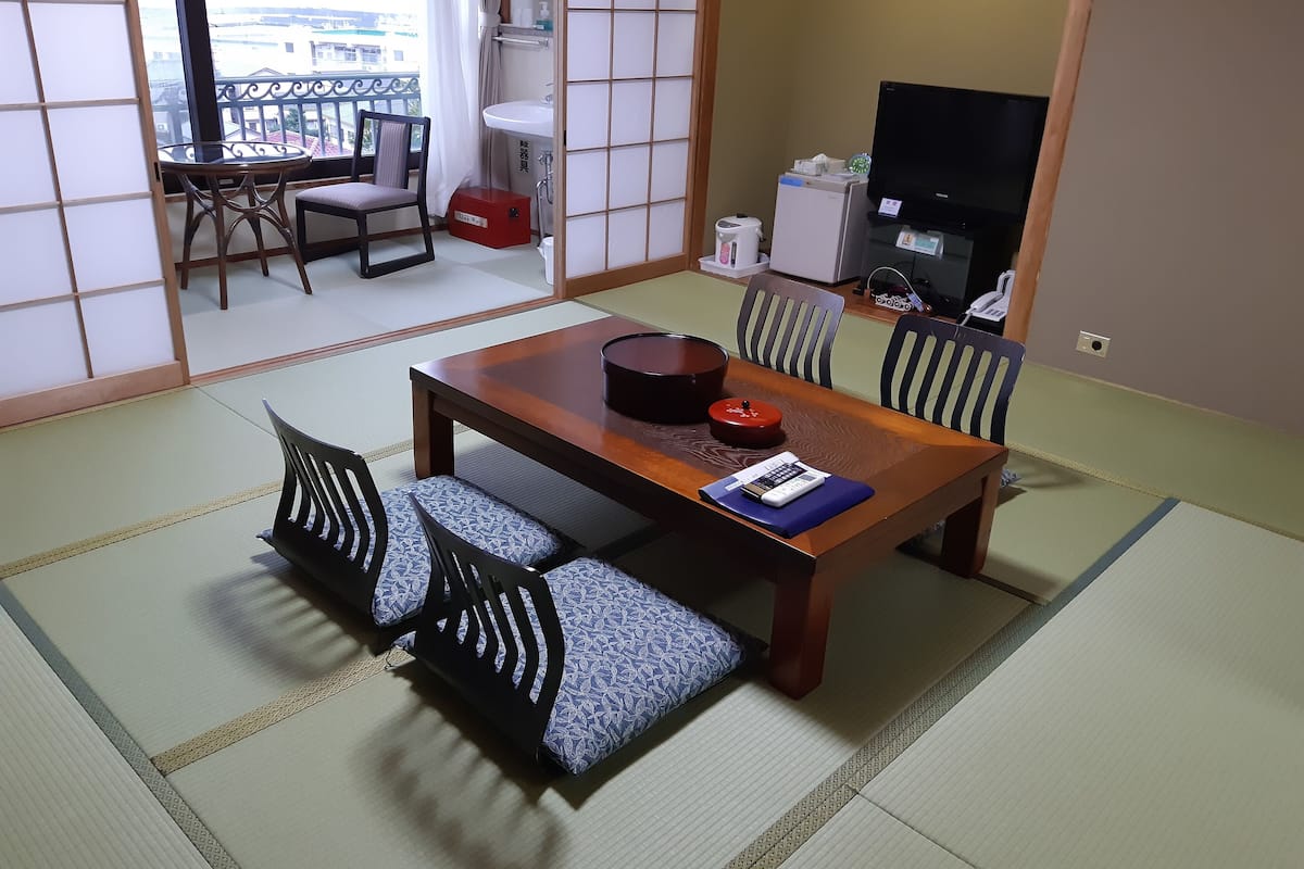 Ein traditioneller japanischer Raum mit Tatami-Boden, einem Holztisch mit Kissen und einem Fernseher.