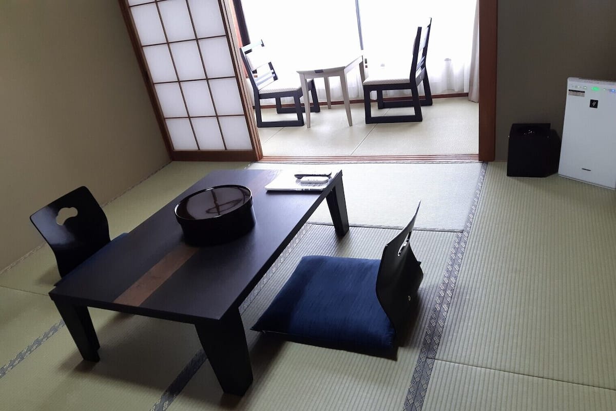 Ein Raum mit Tatami-Matten, einem niedrigen Tisch, zwei Stühlen und einem kleinen Tisch mit einer Tasse, sowie einer Schiebetür.