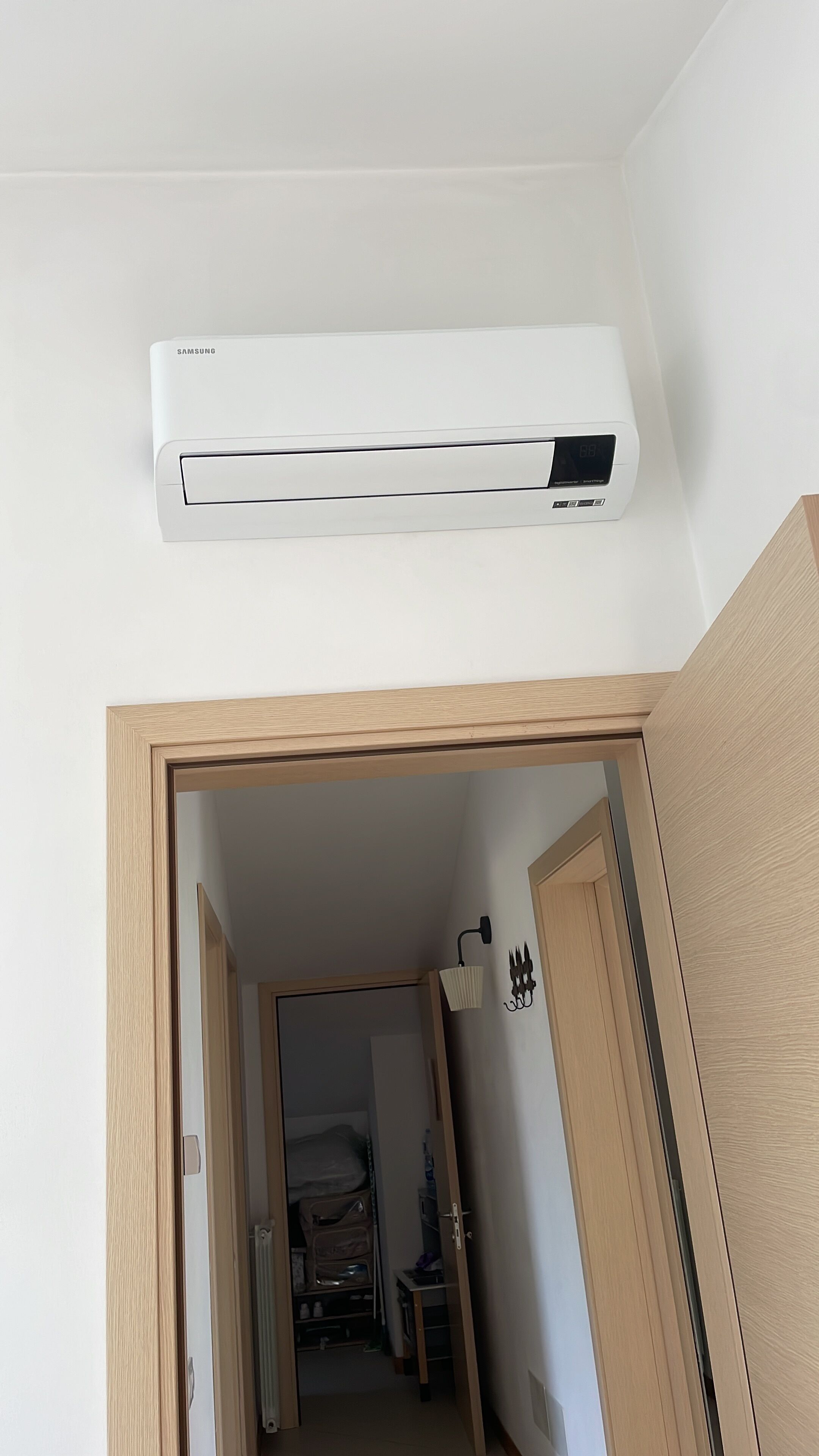 Eine weiße Samsung-Klimaanlage ist an einer Wand in einem Raum angebracht, mit einem Holzrahmen einer Tür und einem kleinen Flur, der zu einem weiteren Raum führt.