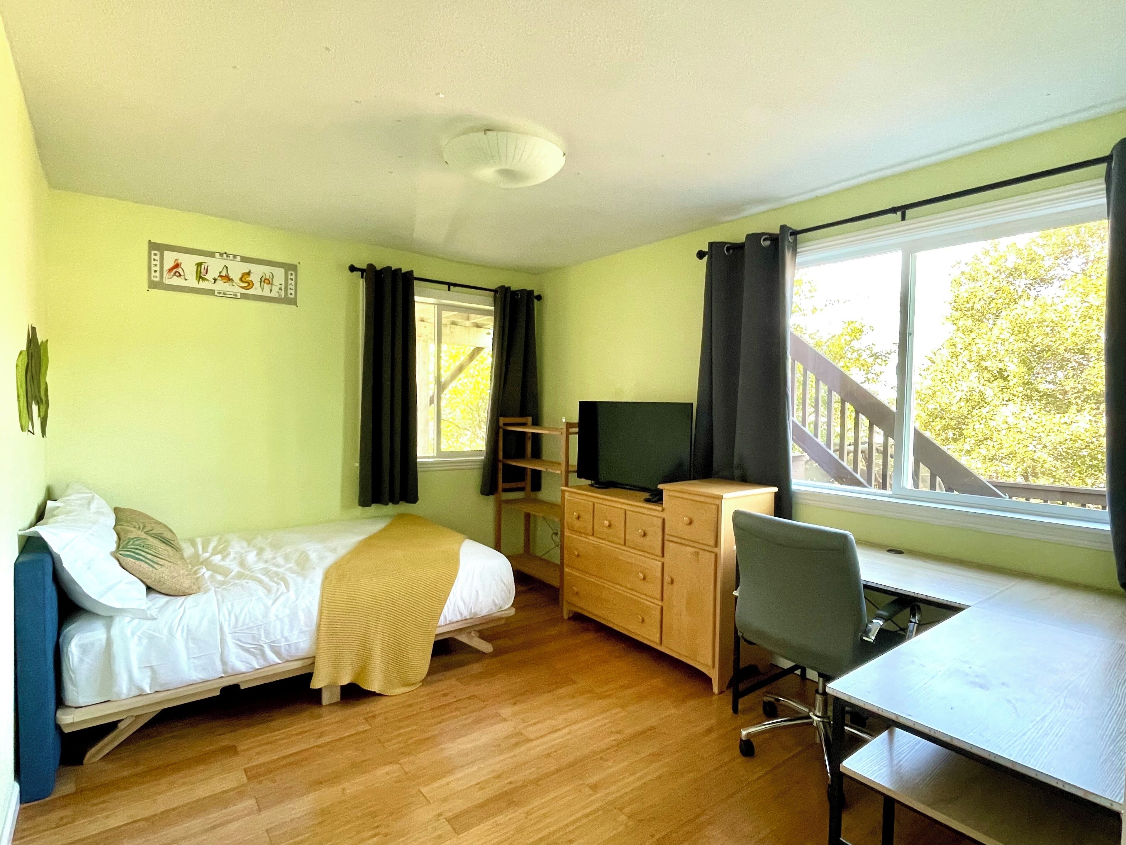 Une chambre à coucher comprenant un lit, un bureau, une chaise, une commode, un téléviseur et une fenêtre avec des rideaux.