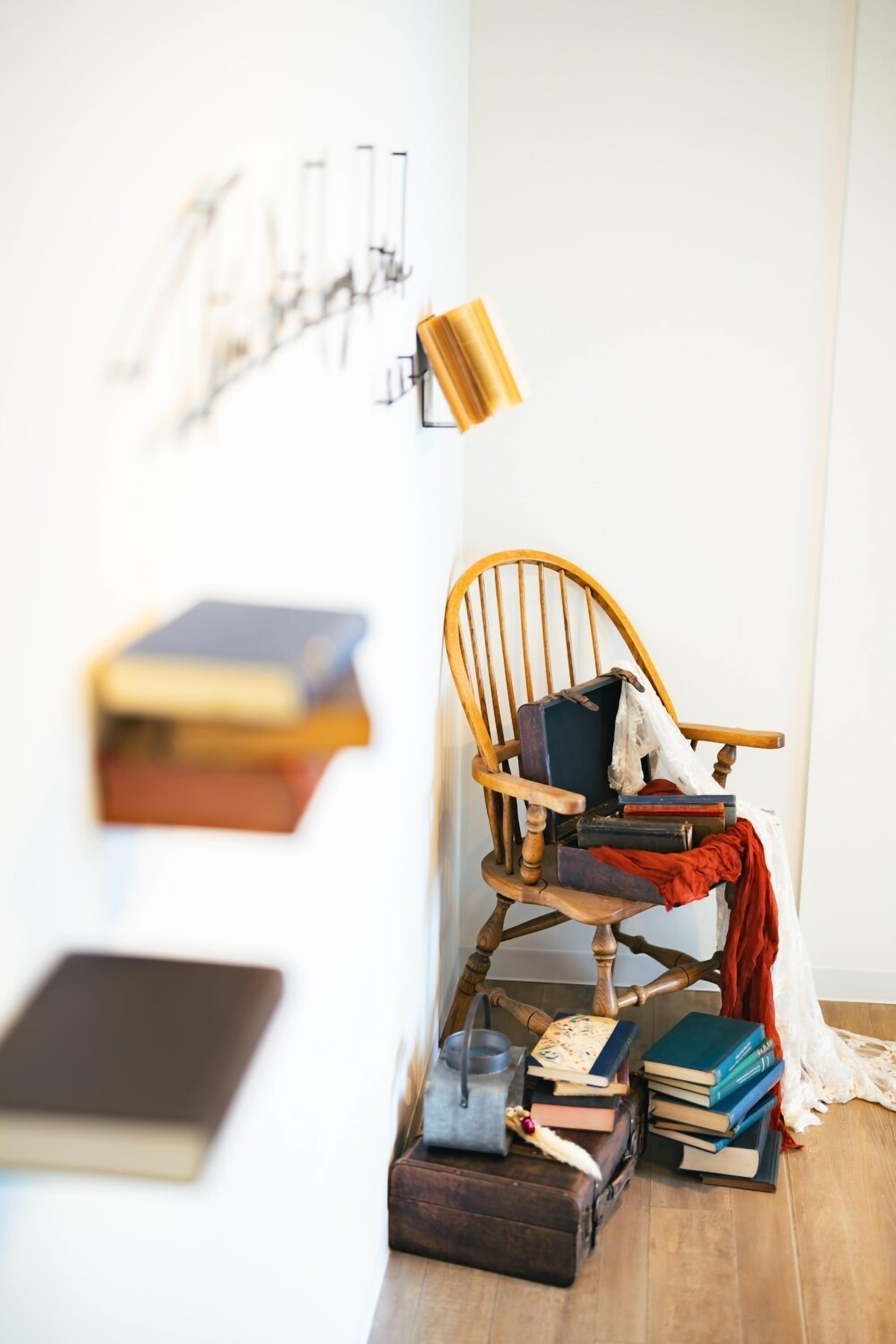 Ein hölzerner Schaukelstuhl mit Büchern und einem roten Stoff, ein kleiner Tisch mit einer Vintage-Lampe sowie eine wandmontierte Leuchte.