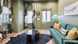 Un salon avec un canapé vert, une table basse ronde et un tapis noir. Il y a deux fenêtres avec des rideaux, une lampe et un tableau encadré au mur.