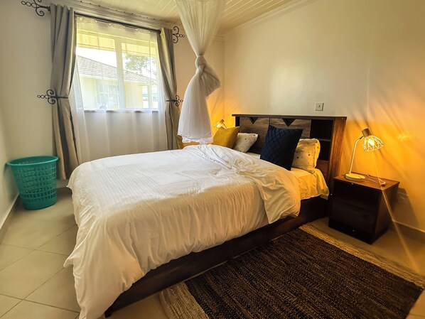 A neatly made bed with white linens, a dark headboard, and a bedside lamp. There is a small trash bin and a window with curtains.