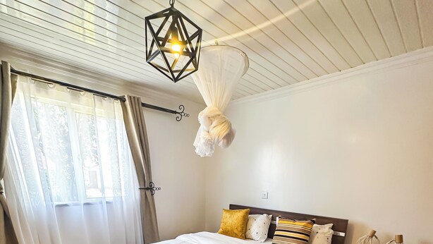 Une chambre à coucher comprenant un lit, des tables de chevet, un luminaire suspendu et une fenêtre avec des rideaux.