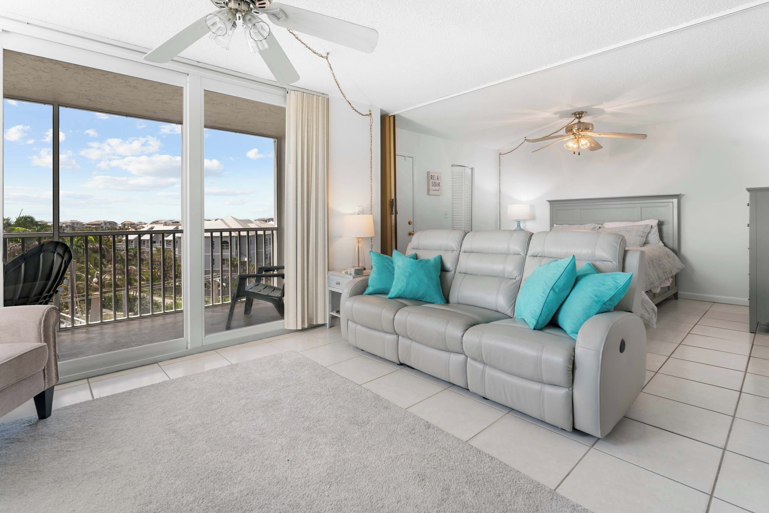 Een moderne woonkamer met een grijze leren bank, teal kussens, een glazen deur naar een balkon en een plafondventilator.