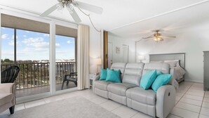 A modern living room with a grey leather sofa, teal cushions, a glass door leading to a balcony, and a ceiling fan. - Gorgeous Beach and Tennis Condo 5608 (Bonita Springs)