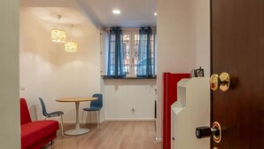 Habitación pequeña con una mesa de comedor, sillas, un sofá rojo, una ventana con cortinas, una puerta y un refrigerador.