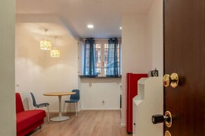 Habitación pequeña con una mesa de comedor, sillas, un sofá rojo, una ventana con cortinas, una puerta y un refrigerador.