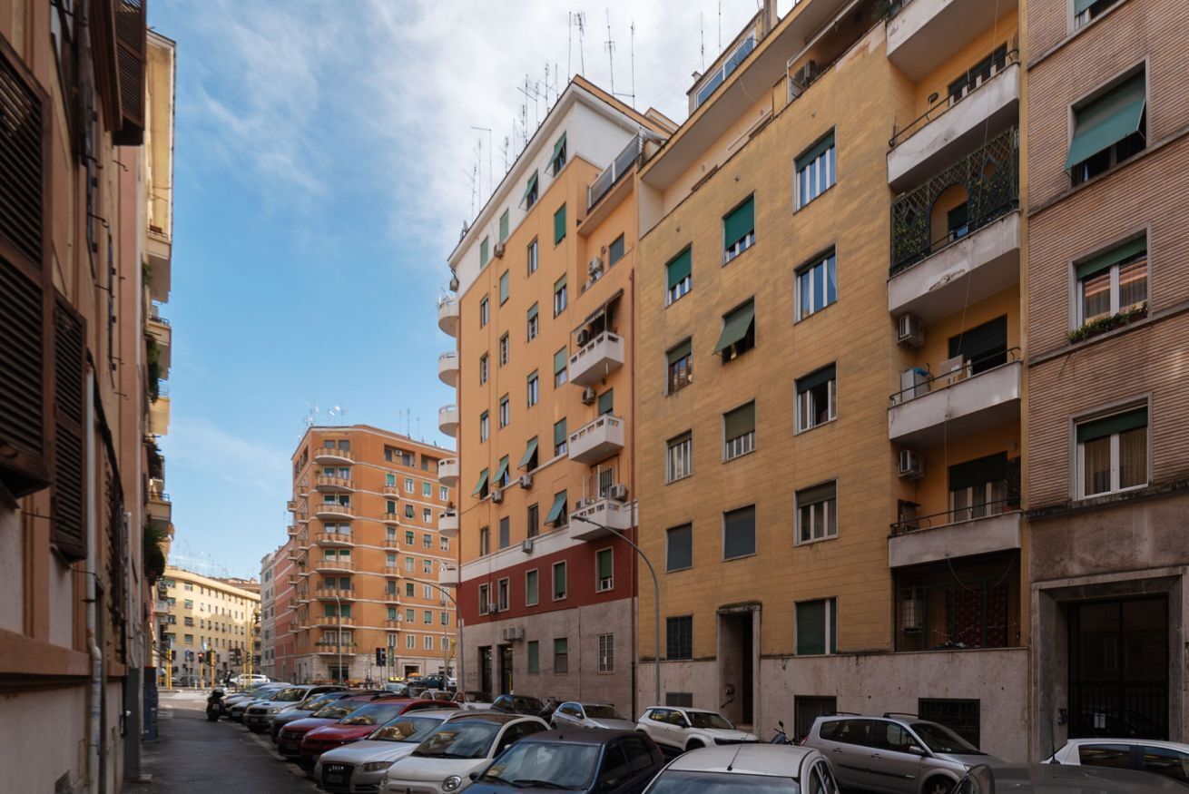 Une rue étroite en milieu urbain, bordée d’immeubles d’habitation à plusieurs étages, de voitures garées et sous un ciel dégagé.