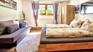 A cozy bedroom with a bed, a sofa, a window with curtains, a radiator, and a TV on the wall.