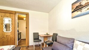 A compact room with a wooden door, a small dining table, two chairs, a sofa, and a bed with a floral bedspread.