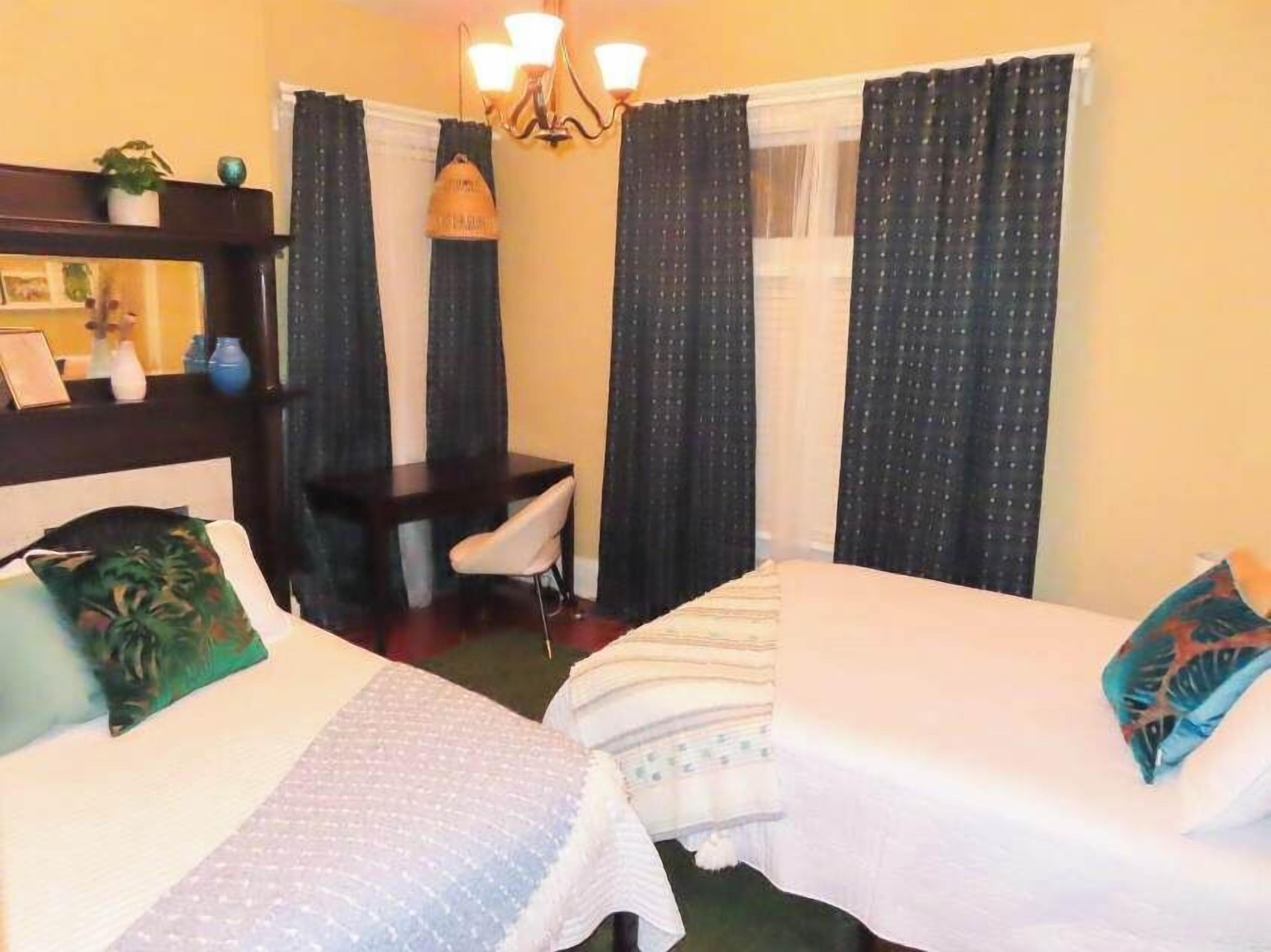 Quarto de hotel com duas camas, uma escrivaninha, uma cadeira, um lustre e uma janela com cortinas.