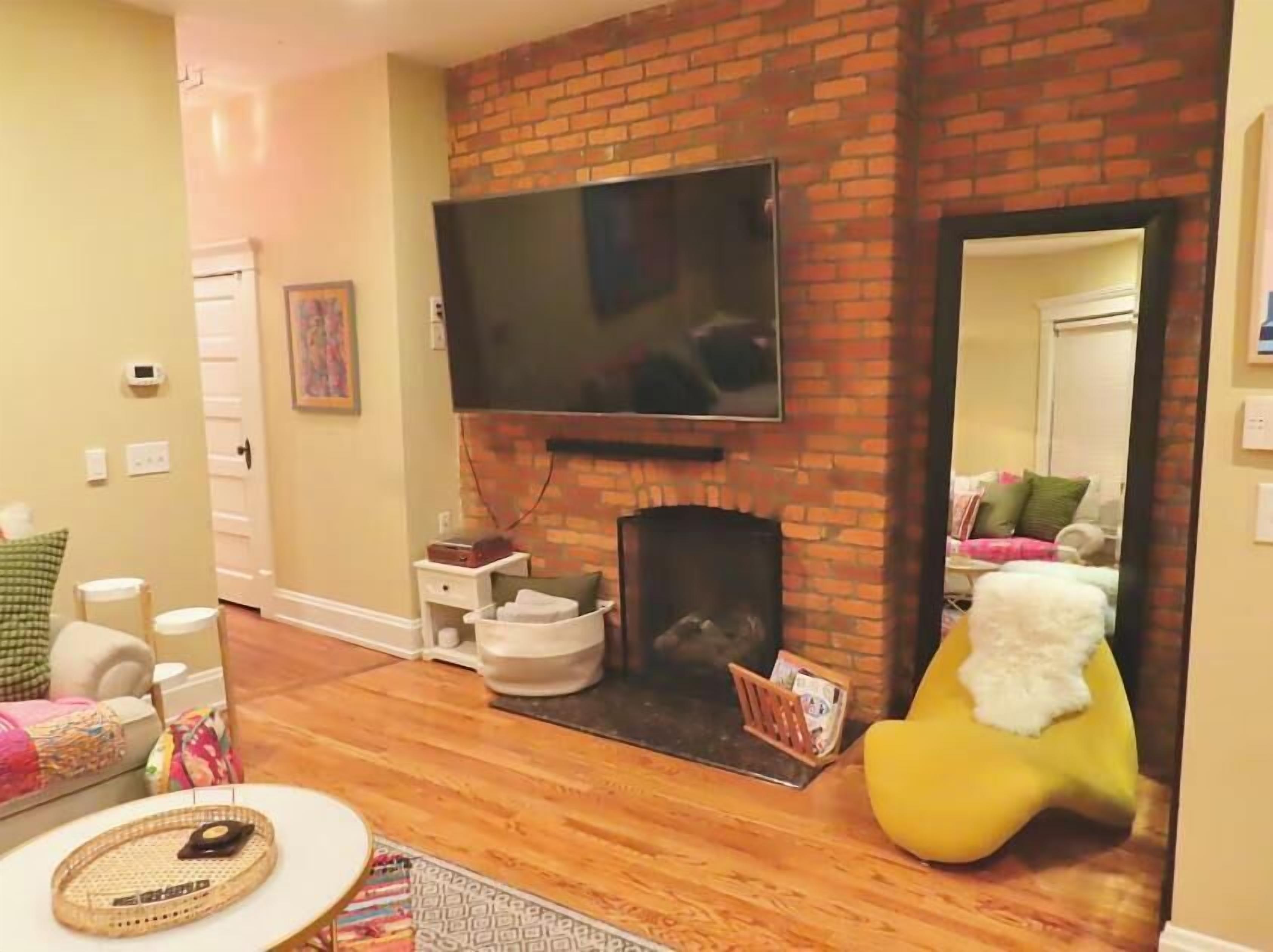 A living room with a brick fireplace, a flat-screen TV mounted above it, a yellow bean bag chair, and a wooden floor.