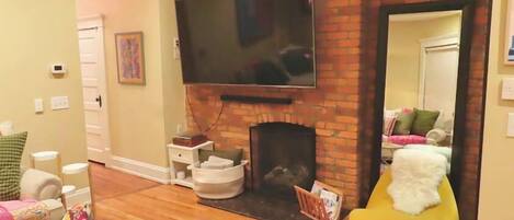 A living room with a brick fireplace, a flat-screen TV mounted above it, a yellow bean bag chair, and a wooden floor.