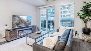 A modern living room with a large flat-screen TV, a leather sofa, a glass coffee table, and a balcony with a view of buildings.