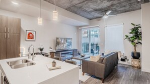 A modern kitchen with a large island, a dining area with a sofa, and a living room with a TV and a plant.