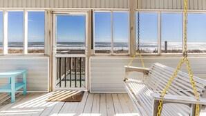 A wooden deck with a white bench and a turquoise table, overlooking a beach with a fence and a clear sky.