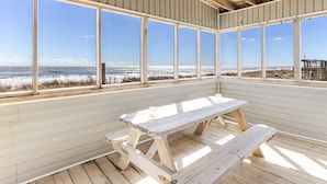 A wooden picnic table in a covered outdoor area with large windows overlooking a beach.