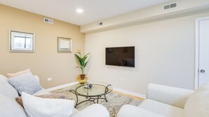 A living room with a glass coffee table, beige walls, a flat-screen TV, and two sofas. - Steps to Shops & Eats: Apt in Portage Park! (Chicago)