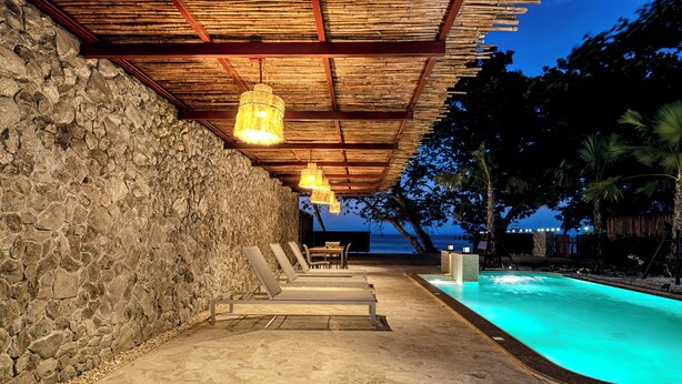Zona de piscina con muros de piedra y vigas de madera, una mesa y sillas, y vista al océano al anochecer.