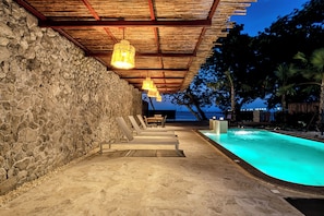 A pool area with stone walls and wooden beams, a table and chairs, and a view of the ocean at dusk.