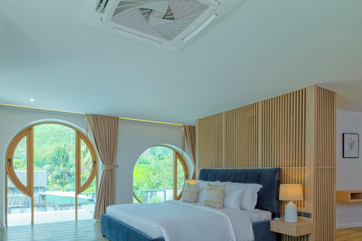 Um quarto espaçoso com uma cama grande, cabeceira de madeira e vista para o verde através de janelas amplas.