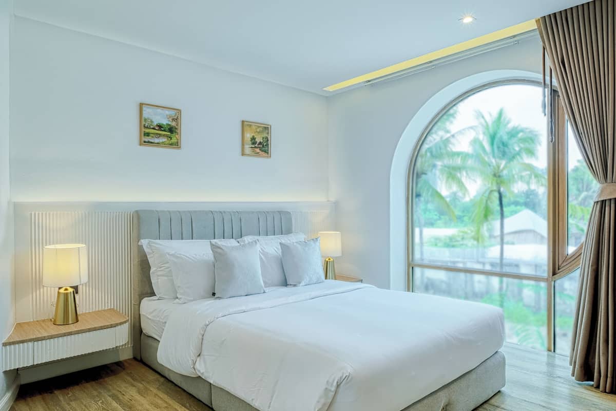 Quarto de hotel moderno com uma cama grande, duas mesas de cabeceira e uma janela ampla com vista para palmeiras.