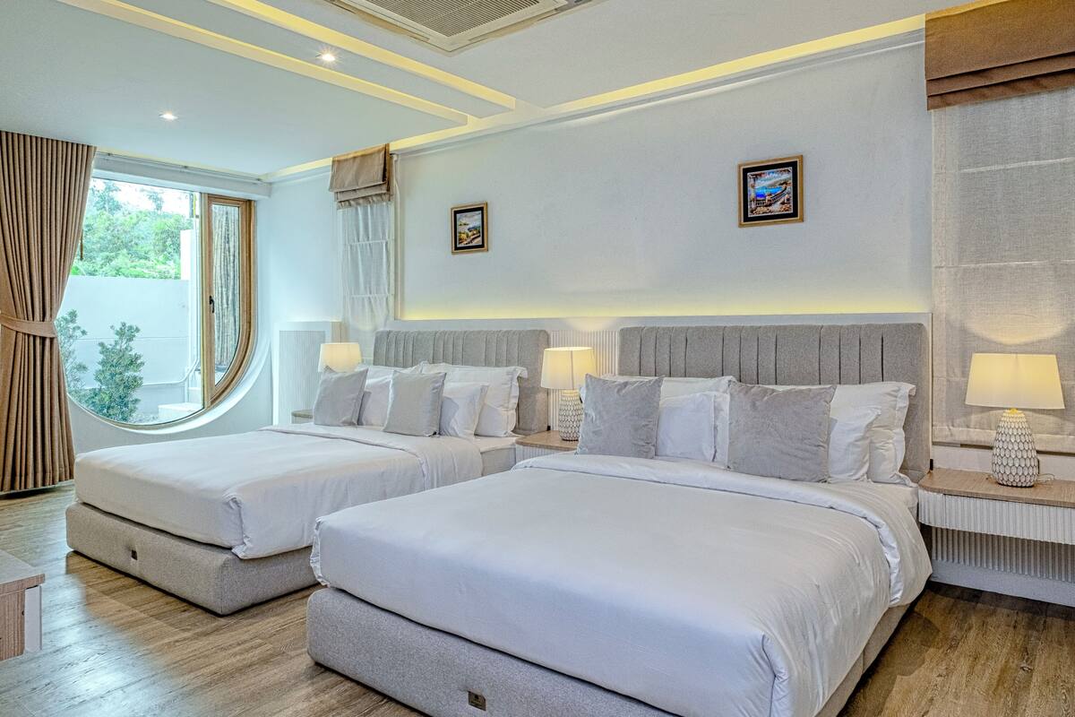 Quarto de hotel moderno com duas camas, uma janela grande e vista para área verde.