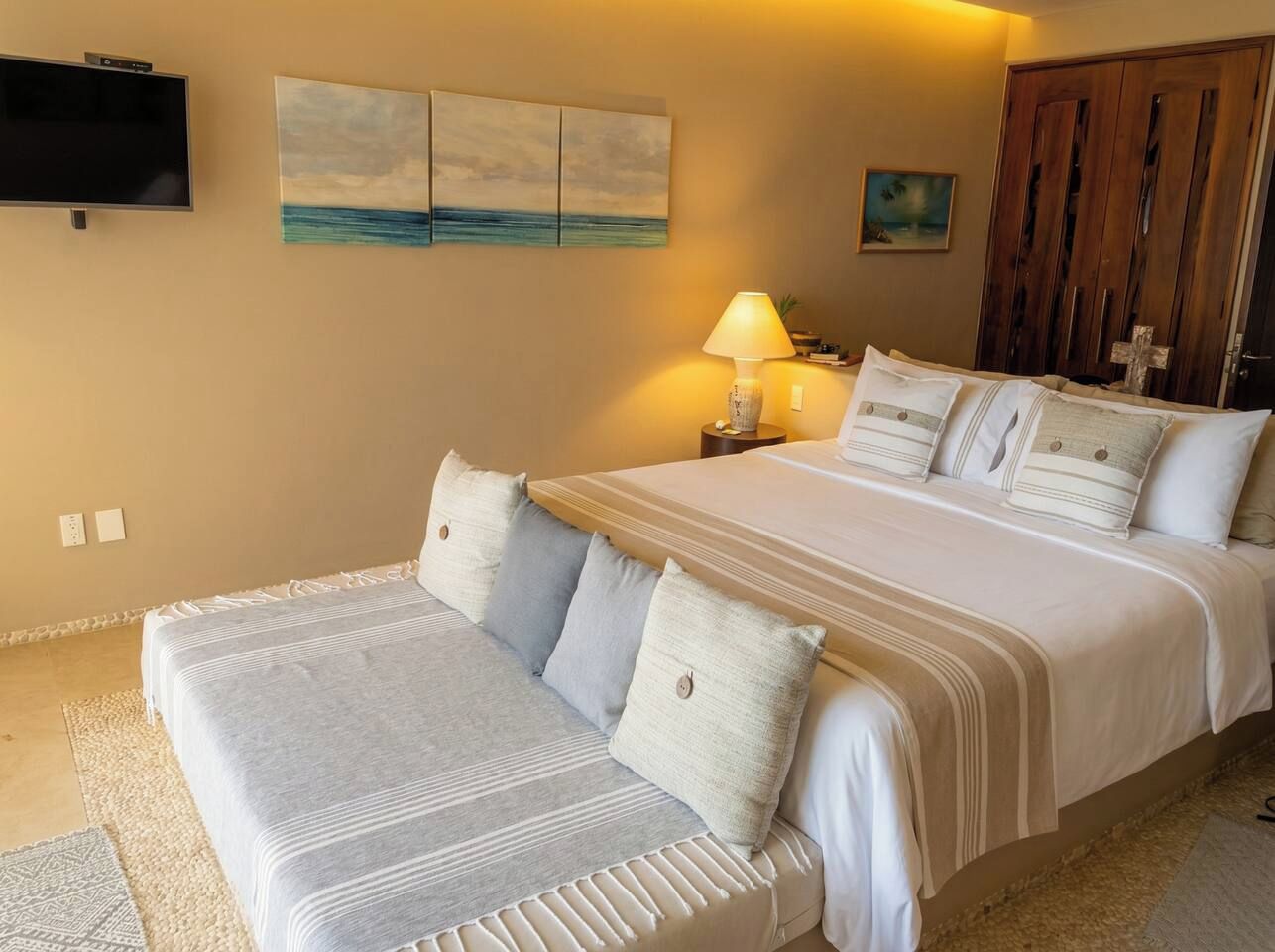Una habitación de hotel con una cama grande, una mesita de noche con lámpara, un televisor montado en la pared y obras de arte en las paredes.