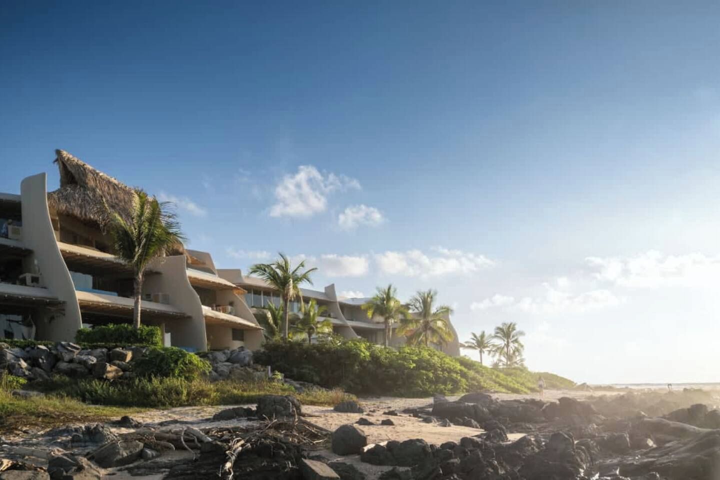 A beachfront hotel with multiple balconies, palm trees, and a rocky shoreline.