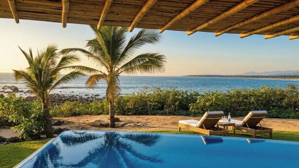 A swimming pool with a view of the sea, palm trees, and lounge chairs.