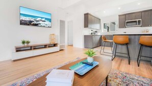 Ein modernes Wohnzimmer mit Couchtisch aus Holz, einem an der Wand montierten Fernseher, einer Küchenzeile mit Mikrowelle und einer Sitzecke mit Barhocker.