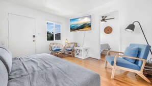 Un dormitorio con una cama, una silla azul, sillones de mimbre, un televisor, un ventilador de techo y una ventana.