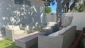 Un patio amueblado con muebles de mimbre, incluyendo sofás y una mesa de centro, rodeado por una cerca blanca y árboles.