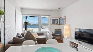 Ein modernes Wohnzimmer mit einem weißen Sofa, einem Esstisch mit blauem Teller, einem Fernseher und Meerblick durch große Fenster.