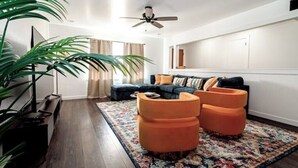 Una sala de estar moderna con piso de madera oscura, una planta grande, un sofá azul con otomanas naranjas y un ventilador de techo.