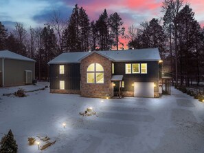 Une maison à deux étages avec un garage, entourée d’un paysage enneigé et d’arbres.