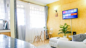 Een moderne woonkamer met een marmeren salontafel, een witte bank, een flatscreen-televisie aan de muur, een eethoek met een kleine tafel en stoelen, en een groot raam met transparante gordijnen.