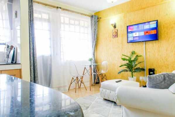 Ein modernes Wohnzimmer mit einem Marmortisch, einem weißen Sofa, einem an der Wand montierten Flachbildfernseher, einem Essbereich mit Tisch und Stühlen sowie einem großen Fenster mit transparenten Vorhängen.
